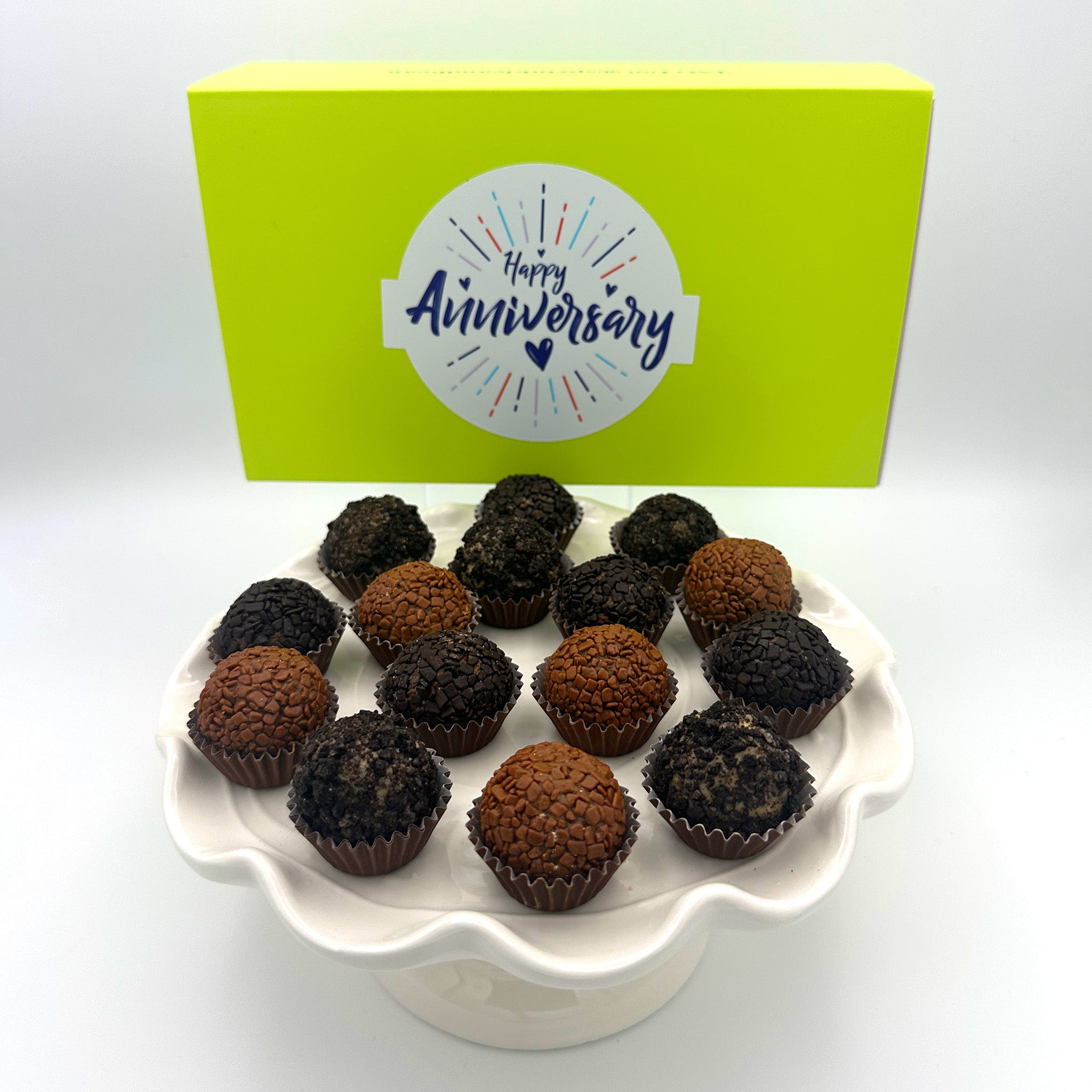 Anniversary Chocolate Lovers Brigadeiros, beloved by chocolate lovers worldwide, are delicately placed on a white plate alongside a charming yellow box, making them a perfect treat for any anniversary celebration.